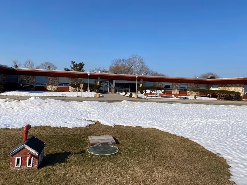 Exterior view of a single-story building with stone and red paneling, surrounded by patches of snow and grass under a clear blue sky. There is a small decorative red and white structure in the foreground on the grass.