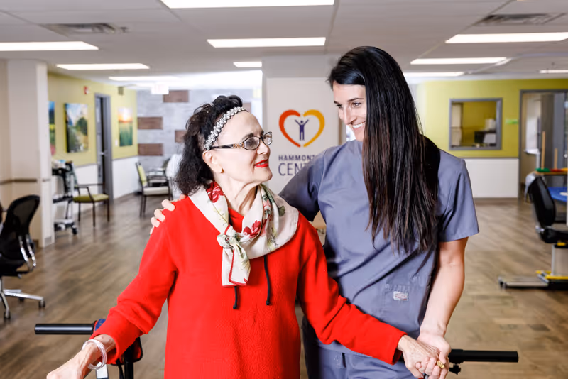 An elderly woman wearing a red sweater and floral scarf is being assisted by a female caregiver in gray scrubs inside a bright, spacious room with wooden flooring and chairs. The caregiver is smiling and holding the woman's hand, providing support as they walk together. In the background, there is a wall with the Hammonton Center logo featuring a heart and a person figure.
