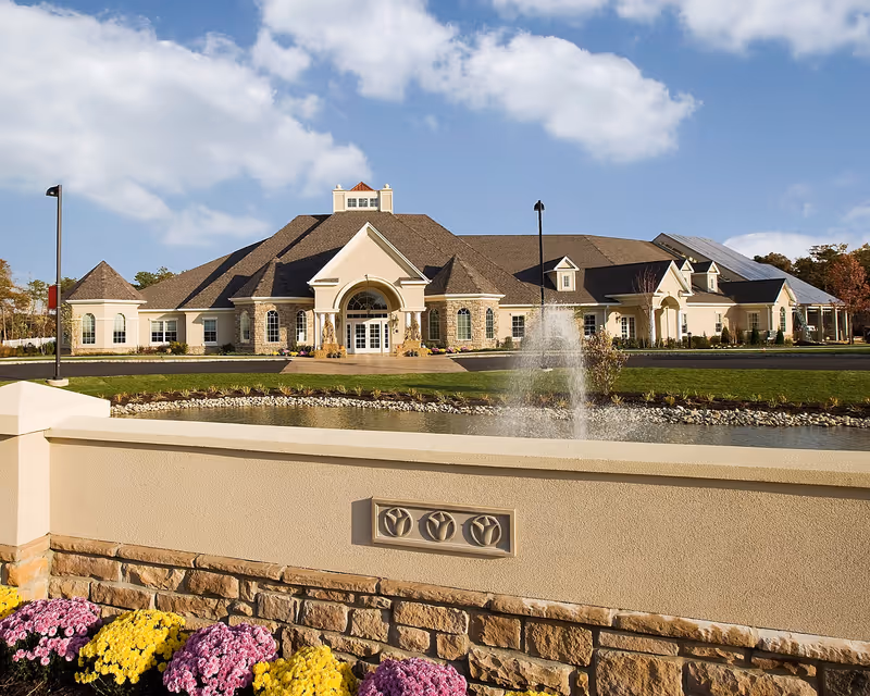 Front exterior of a large senior living facility with a fountain, manicured lawn, and colorful flower beds under a blue sky.