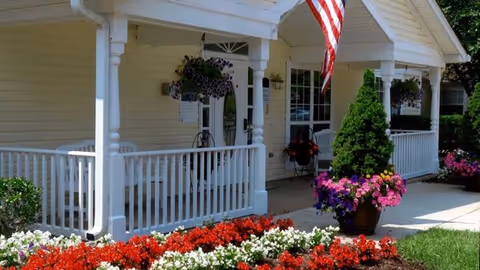 Front porch of a light yellow building with white railings and columns, decorated with hanging flower baskets and potted plants. An American flag is displayed near the entrance, and colorful flowers are planted in front of the porch.