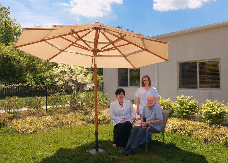 An elderly man sitting on a chair outside under a large beige patio umbrella with two female healthcare workers standing and sitting beside him. They are in a garden area with green grass, bushes, and a building with windows in the background under a partly cloudy blue sky.