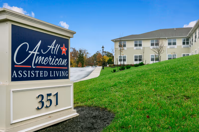 Exterior view of All American Assisted Living at Washington Township showing a large sign with the facility name and address number 351, with a green grassy hill and a multi-story building in the background under a blue sky with some clouds.