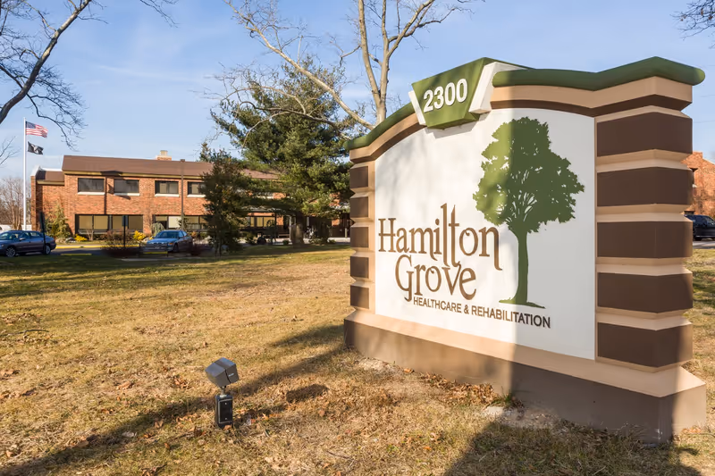 Outdoor view of Hamilton Grove Healthcare and Rehabilitation facility sign with the building and parked cars in the background under a clear sky.