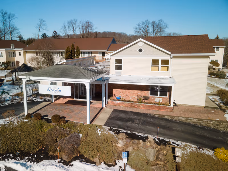Front exterior of a two-story nursing facility with a covered entrance/porte-cochere and a sign reading "Boonton" amid a light snowy landscape.