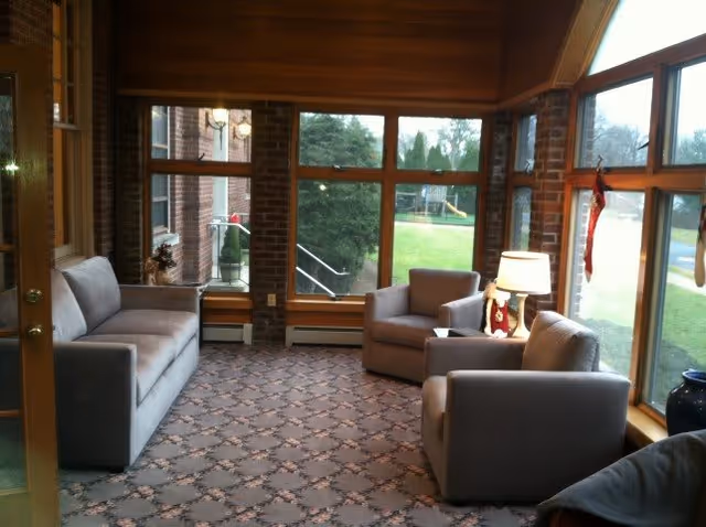 A cozy sitting area with a gray sofa and two matching armchairs arranged around a small side table with a lamp. The room has large windows letting in natural light and offering a view of a green lawn and playground outside. The walls are brick and wood-paneled, and the floor is carpeted with a patterned design.