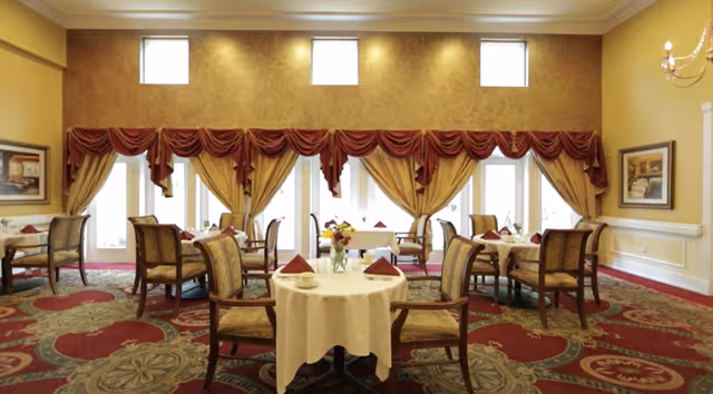 A dining room with round tables covered in white tablecloths, each set with cups, plates, and folded napkins. The room has large windows with gold curtains and red valances, ornate patterned carpet, and framed artwork on the walls.