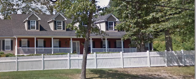 Exterior view of a single-story residential building with a gray roof, multiple dormer windows, a front porch with white railings, and a white picket fence in front. Trees are visible around the building.
