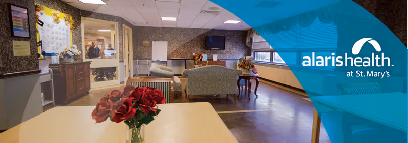 Interior view of a senior living facility lounge area with comfortable seating including armchairs and a sofa, a television mounted on the wall, and a table with a vase of red roses in the foreground. The room has large windows allowing natural light and a patterned wallpaper. A hallway and reception area are visible in the background.