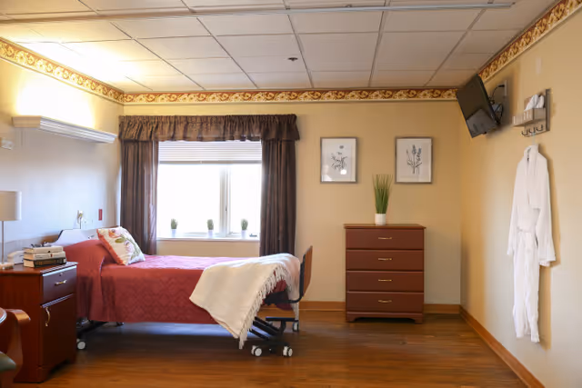 A cozy bedroom in a senior living facility with a single bed covered in a red quilt and a white blanket draped over the foot. There is a wooden nightstand with books and a lamp next to the bed. A window with dark curtains and small potted plants on the sill lets in natural light. On the opposite wall, there is a wooden chest of drawers with a potted plant on top and two framed botanical prints hanging above. A white robe hangs on the wall near a mounted television.
