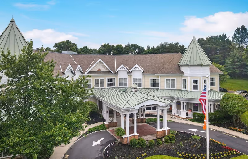 Front exterior of a two-story senior living facility with a covered entrance, landscaped driveway, and an American flag.