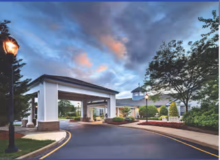 Covered porte-cochère and circular driveway at the front of a landscaped senior living facility under a dramatic cloudy sky.