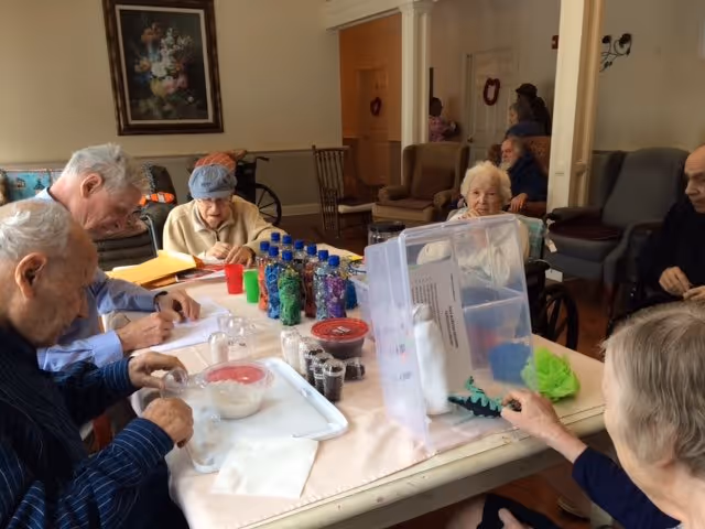 A group of elderly individuals seated around a table engaged in a craft or activity session in a common room. The table is covered with various craft supplies including bottles filled with colorful materials, small containers, and papers. The room has chairs and a painting on the wall in the background.
