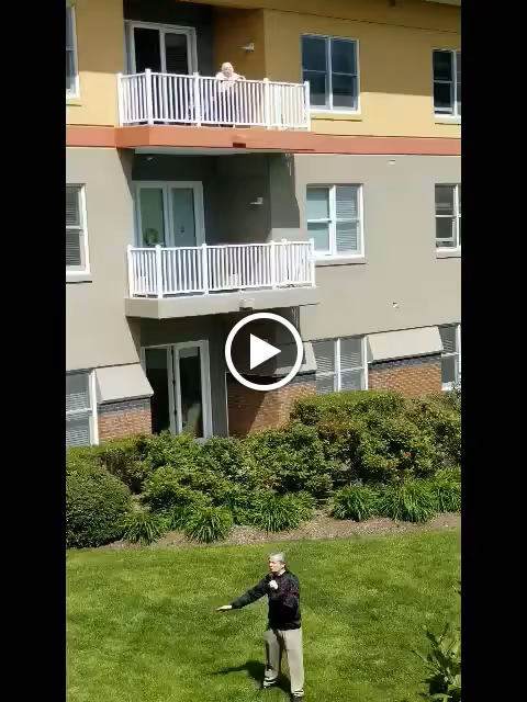 A man standing on a grassy lawn in front of a multi-story residential building with balconies and windows. The building has beige and light brown exterior walls with some brickwork at the bottom. There are bushes and plants along the edge of the lawn.