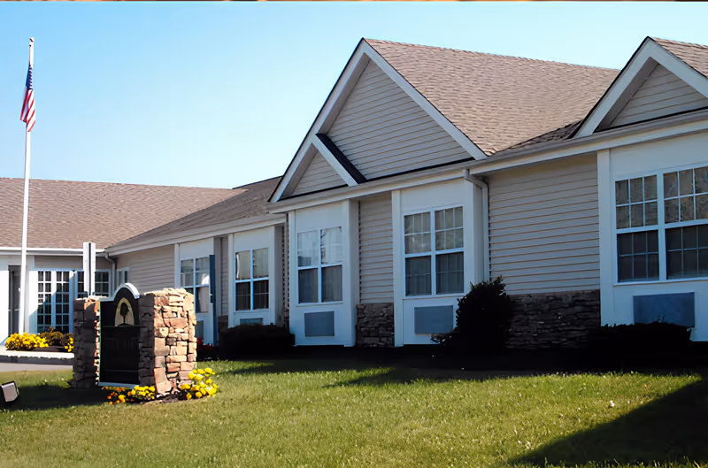 Front exterior of a single-story assisted living building with beige siding, multiple windows, a flagpole, and a landscaped lawn.