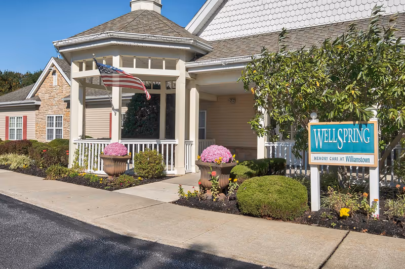 Front entrance of a senior living facility with a covered porch, American flag, potted flowers, landscaped shrubs, and a sign reading 'WELLSPRING'.