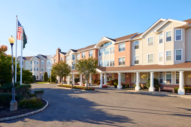 Exterior view of a senior living facility building with a driveway, landscaped garden beds, and three flagpoles displaying the American flag and other flags under a clear blue sky.