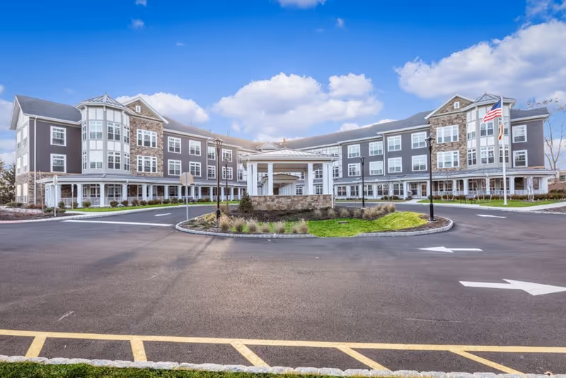 Front view of a three-story senior living facility with a covered entrance, circular driveway, and an American flag.