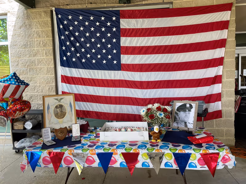 A patriotic display table set up outdoors in front of a large American flag hanging on a brick wall. The table is covered with a colorful balloon-patterned tablecloth and decorated with red, white, blue, and silver triangular pennants. On the table are framed photographs of military personnel, medals, a bouquet of red, white, and blue flowers, an eagle figurine, and a box of decorated cookies. To the left, there are star-shaped balloons with American flag designs.