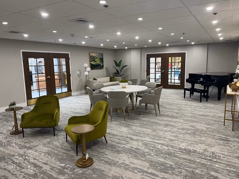 Spacious senior living common room with green lounge chairs, a round table with chairs, and a black grand piano near double French doors.