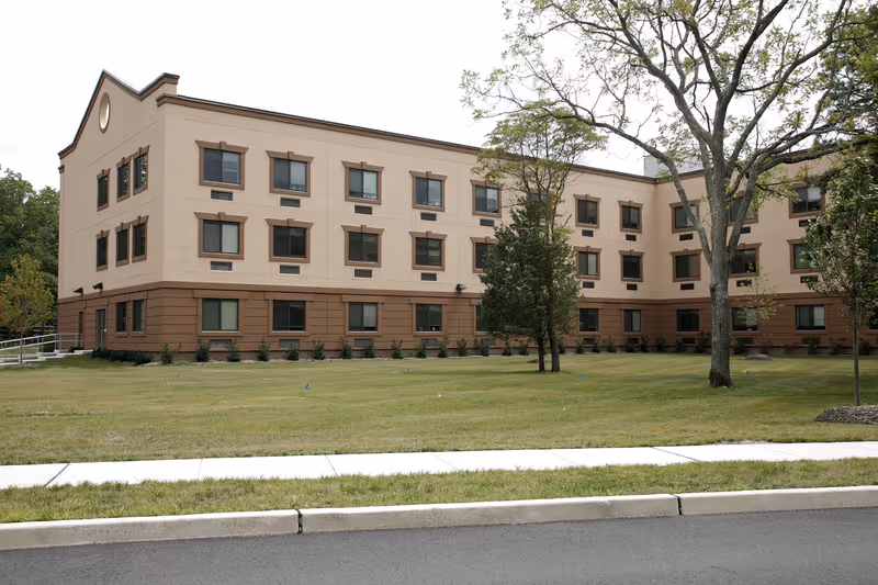 Exterior view of a three-story beige and brown building with multiple windows, surrounded by a grassy lawn with a few trees and a sidewalk in the foreground.