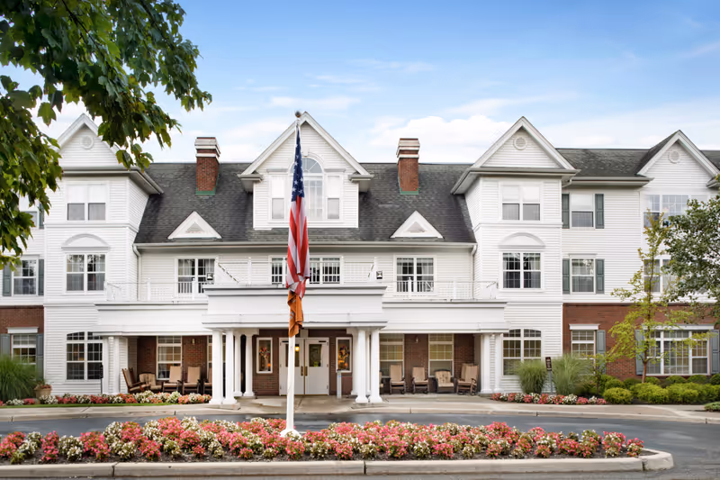 Front exterior of a three-story white senior living building with a covered porch entrance, an American flag, and flowerbeds.