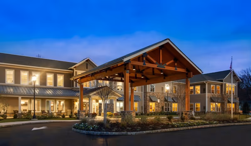 Exterior view of Senior Living Residences Marlboro building at dusk with lights on inside. The building features a large covered entrance with wooden beams and stone pillars, surrounded by landscaped grounds and a driveway with a white directional arrow.