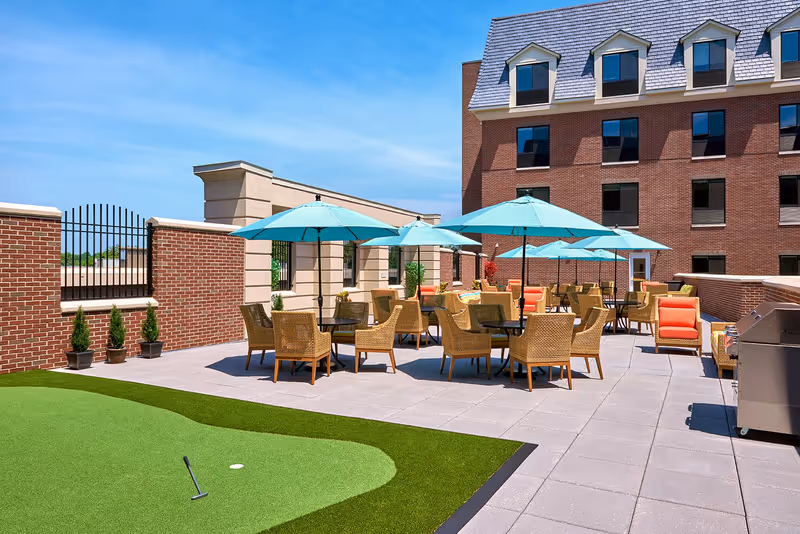 Rooftop patio with wicker tables and turquoise umbrellas, lounge chairs, and a small putting green next to a brick building.