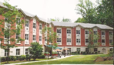 Exterior view of a three-story brick and beige senior living facility building with multiple windows, surrounded by green trees and a well-maintained lawn with a patio area featuring tables and umbrellas.