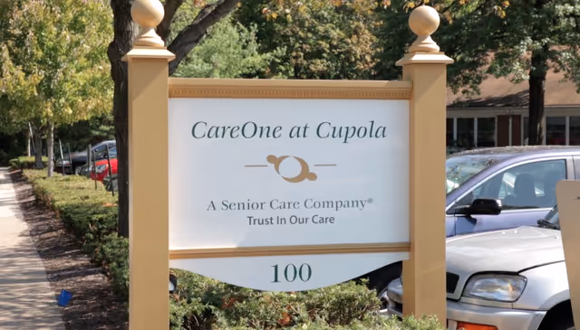 Entrance sign reading "CareOne at Cupola — A Senior Care Company, Trust In Our Care, 100" set among landscaping with parked cars and trees behind it.