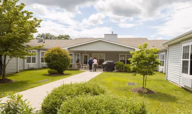 Sunny courtyard with a paved walkway leading to the front of a single-story residential building, landscaped lawns and shrubs, and two people near the entrance.