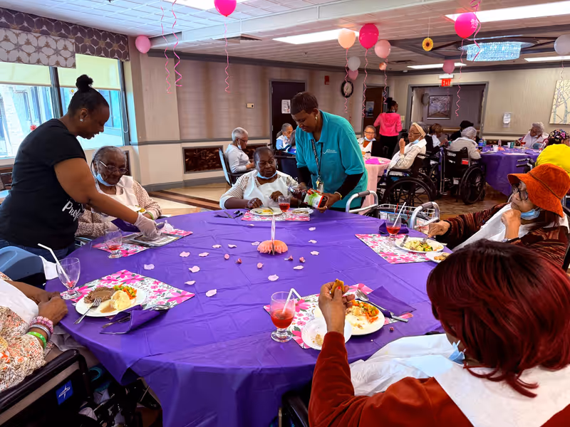 A group of elderly people seated around a large round table covered with a purple tablecloth in a decorated dining room. Two caregivers are assisting them, one serving food and the other pouring a drink. The room is decorated with pink and red balloons hanging from the ceiling, and the elderly individuals are enjoying a meal together.