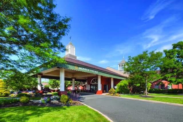 Exterior view of Monroe Village senior living facility entrance with a covered driveway, surrounded by green trees and landscaped gardens under a clear blue sky.
