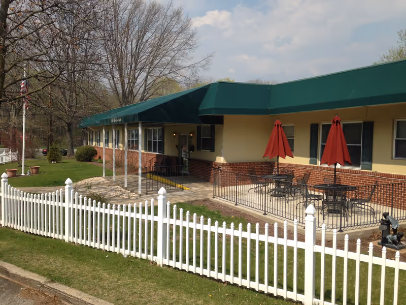 Exterior view of a single-story building with a green awning and a brick lower facade. There is a white picket fence in the foreground, a small patio area with tables and red umbrellas, and a flagpole with an American flag. Trees without leaves are visible in the background under a partly cloudy sky.