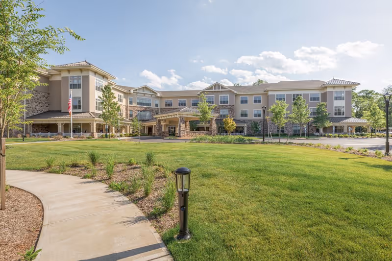 Exterior view of Sunrise of Bridgewater senior living facility showing a large, three-story building with a covered entrance, surrounded by well-maintained green lawns, young trees, and a curved concrete walkway under a partly cloudy sky.