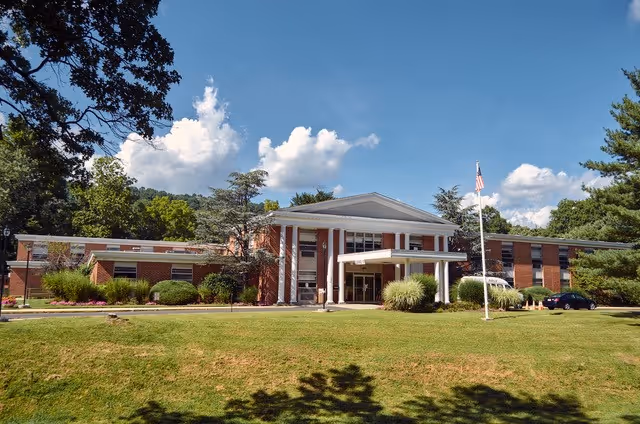 Front exterior view of a single-story brick building with white columns at the entrance, surrounded by green grass, trees, and a clear blue sky with clouds. An American flag is flying on a flagpole near the entrance.