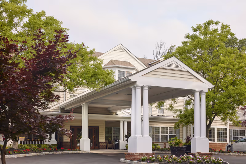 Front exterior view of a senior living facility with a covered entrance supported by white columns, surrounded by green trees and landscaped flower beds.