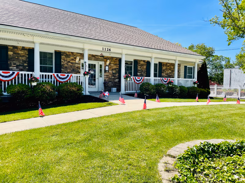 Front exterior view of a single-story building with stone and brick facade, white columns, and a porch decorated with red, white, and blue bunting and small American flags lining the walkway. The building number 1126 is visible above the entrance door. The lawn is green and well-maintained with a circular garden bed in the foreground.