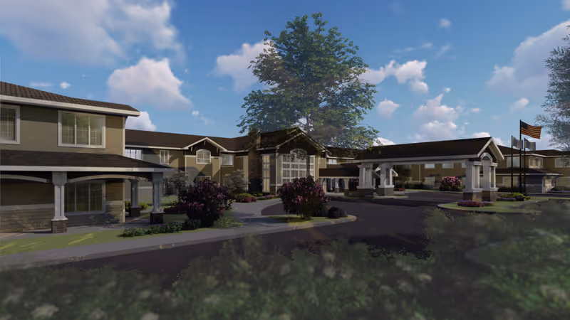 Exterior view of Juniper Village at Hamilton, showing a large building with multiple windows, a covered entrance with pillars, landscaped bushes and trees, and an American flag on a flagpole under a partly cloudy blue sky.