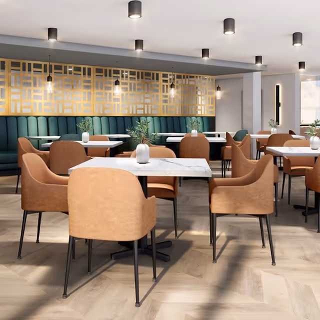 A modern dining room with multiple square white marble tables surrounded by tan upholstered chairs. The back wall features a green cushioned bench with a decorative gold geometric panel above it. Small vases with green plants are placed on each table. The room is well-lit with ceiling lights and pendant lights hanging above the bench.