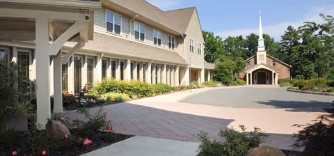 Exterior view of a senior living facility with a large paved courtyard, beige multi-story building on the left, and a small chapel with a white steeple on the right, surrounded by greenery and clear blue sky.