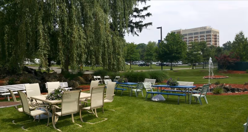 Outdoor seating area with multiple tables and chairs on a grassy lawn, surrounded by trees and landscaping. A fountain is visible in the background along with a large building and parked cars.
