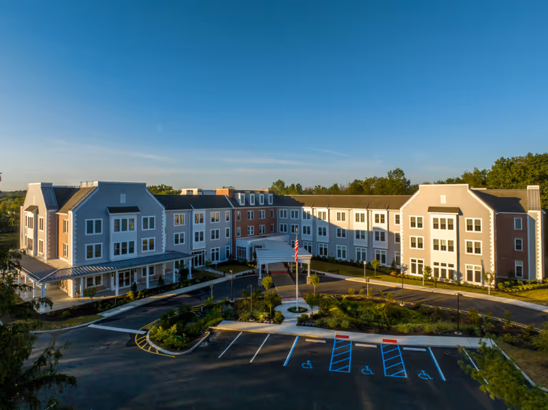 Front exterior of a multi-story senior living building with a circular driveway, flagpole, and parking lot under a clear blue sky.