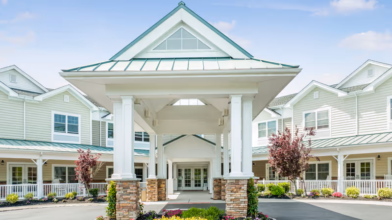 Front exterior view of The Terraces at Seacrest Village senior living facility with a covered entrance supported by white columns and stone bases, surrounded by well-maintained landscaping and a clear blue sky.