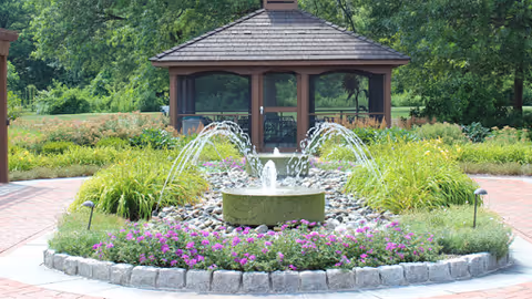 Circular stone fountain with arcing water jets and purple flowers in front of a wooden gazebo in a landscaped garden.
