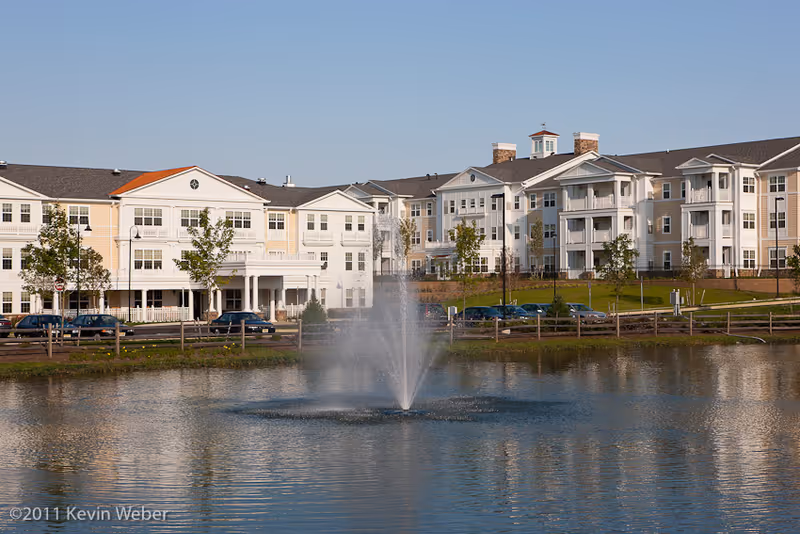 Multi-story senior living building with balconies seen across a pond with a central fountain.