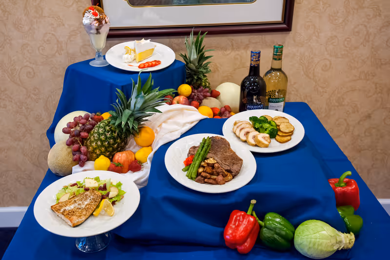 A display of various plated meals and fresh fruits on a blue tablecloth. The dishes include a steak with asparagus and potatoes, sliced chicken with broccoli and roasted potatoes, a piece of cooked fish on a bed of salad, and a slice of lemon pie with whipped cream and strawberries. Surrounding the plates are fresh fruits such as pineapples, grapes, cantaloupe, apples, and strawberries, along with red and green bell peppers and a cabbage. Two bottles of wine are also visible in the background.