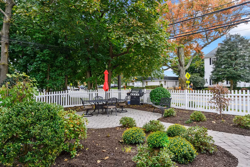 Outdoor patio area with a round table and chairs, a red umbrella, a barbecue grill, and a bench surrounded by green bushes and trees, enclosed by a white picket fence.