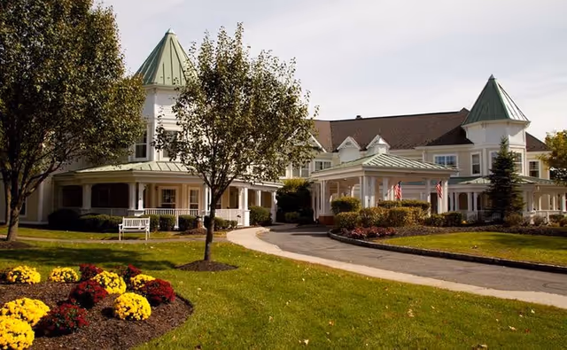 Exterior view of a senior living facility with white buildings featuring green roofs and turrets, surrounded by well-maintained lawns, trees, and flower beds with yellow and red flowers under a clear sky.