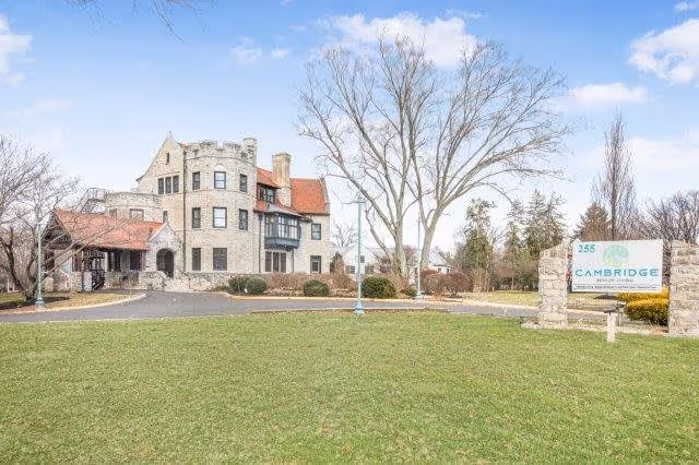 Exterior view of a large stone building with castle-like architectural features, including a turret and red roof tiles, situated behind a well-maintained lawn and trees. A sign in the foreground reads 'Cambridge Enhanced Senior Living'.