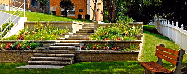 Outdoor garden area with tiered stone steps and retaining walls, landscaped with various plants and flowers. A wooden bench is positioned on the grass near a white picket fence. A brick building and trees are visible in the background.